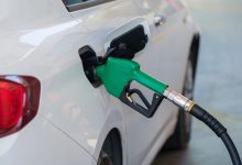 Close de um bico verde abastecendo um carro branco em um posto de combustíveis.