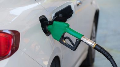 Close de um bico verde abastecendo um carro branco em um posto de combustíveis.