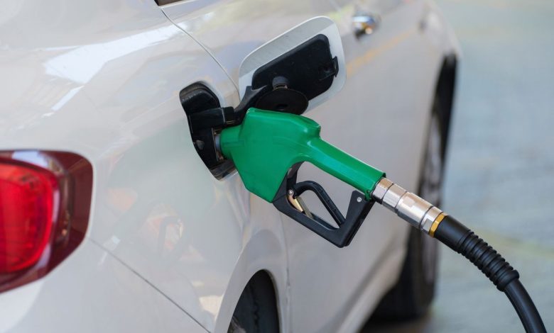 Close de um bico verde abastecendo um carro branco em um posto de combustíveis.